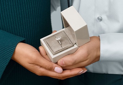 Hands holding an open jewelry box with a completed diamond engagement ring.