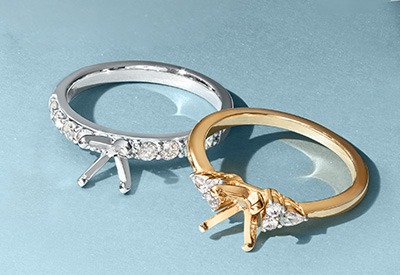 White gold and yellow gold engagement ring settings displayed side by side on a light blue background.