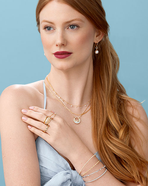 Layered Gold Jewelry and Pearl Earrings Collection Woman wearing layered gold necklaces, pearl drop earrings, rings, and diamond bracelets against a light blue background